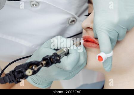Woman having permanent makeup on lips in beauty salon. Cosmetologist in gloves applying tattoo with machine, macro view Stock Photo