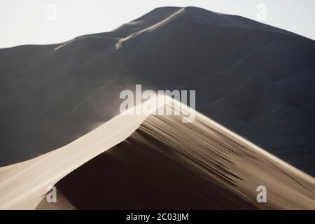 A strong wind lifts sand over the top of a large dune. Sand dunes in the Sahara desert, North Africa. Stock Photo