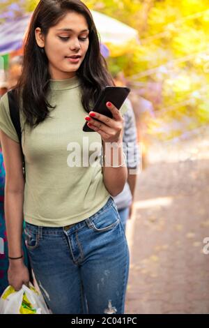 Beautiful happy late teen girl holding shopping bag, walking on market street and using smartphone at day time. Stock Photo
