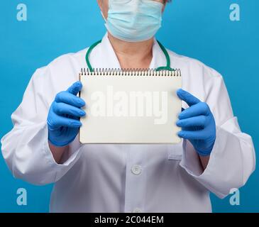 medic woman in white coat, blue latex gloves holding an open notebook with white sheets, blue background, copy space Stock Photo