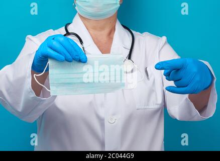 doctor in a white coat and mask holds a stack of protective disposable face masks, physician stands on a blue background