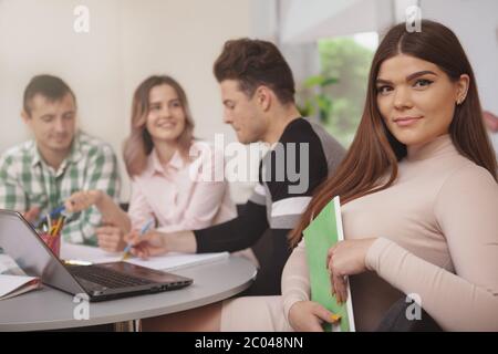 Beautiful young female student smiling to the camera, her classmates working on the background, copy space. Cheerful lovely woman enjoying working on Stock Photo