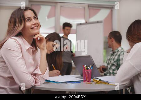 Beautiful happy young female student looking away dreamily, her classmates discussing study project on the background. College students during project Stock Photo