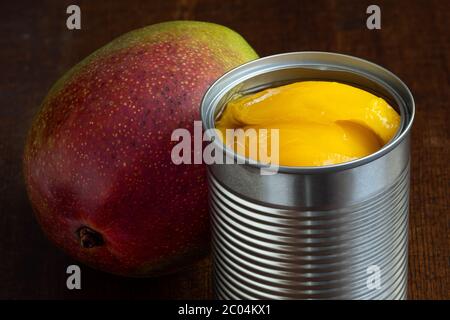 Canned mango slices in syrup in a metal can next to a whole fresh mango isolated on white. Dark wood background. Stock Photo