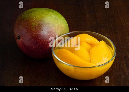 Canned mango slices in syrup in a glass bowl next to a whole fresh mango isolated on white. Dark wood background. Stock Photo