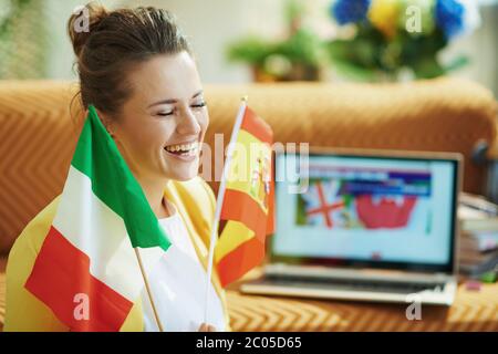 happy trendy 40 years old woman in yellow jacket with flags learning foreign language online on a laptop in the modern living room in sunny day. Stock Photo