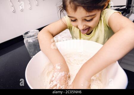 funny little girl with her tongue out mixing flour in a bowl to make homemade bread dough, healthy food concept at home Stock Photo