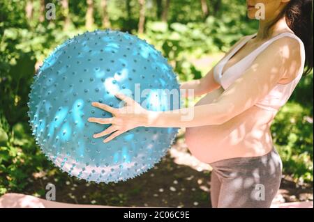 Beautiful young pregnant woman doing yoga exercising with fitness pilates ball in park outdoor. Standing and holding with both hands. Active future Stock Photo