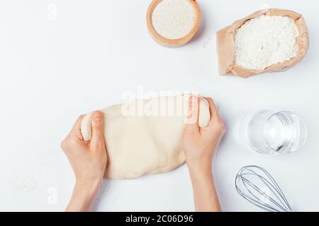 Flat lay female hands kneading dough on white table next to flour, glass of water and whisk. Stock Photo