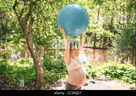 Beautiful young pregnant woman doing yoga exercising with fitness pilates ball in park outdoor. Standing and holding with both hands. Active future Stock Photo