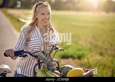 Woman using her bicycle to buy fresh produce standing on a rural road backlit by a warm glow of the sun smiling happily at the camera as she holds her Stock Photo