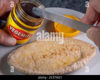 One hand on a jar of newly opened Vegetmite yeast extract, with the other holding a knife above a buttered wholemeal pitta bread on a plate below. Stock Photo