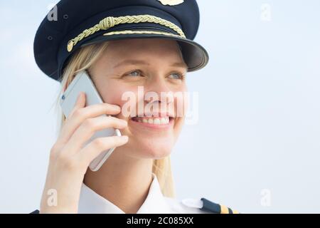 A blonde pretty female pilot talking on her phone at work in the airport outside. Blue sku background. Stock Photo
