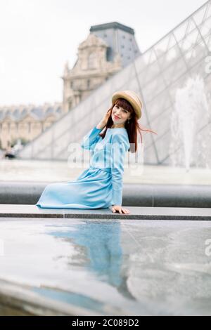 PARIS, FRANCE - September 17, 2019: Young happy woman in blue dress, sitting near the fountain and posing for a photo with the glass pyramid Stock Photo