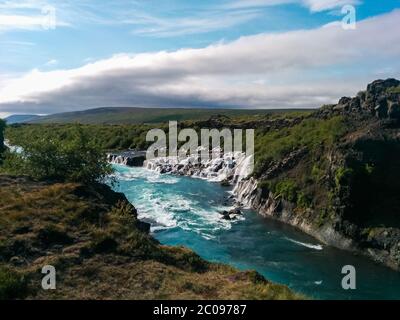 View of Hraunfossar waterfalls in Iceland. Crystal clear beautiful blue waters, green fields and trees on a sunny day. Stock Photo