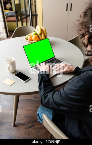 back view of freelancer using laptop with green screen near smartphone, coffee cup and bowl with fresh fruits Stock Photo