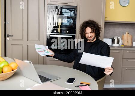 Selective focus of confused freelancer holding papers with charts near gadgets on kitchen table Stock Photo