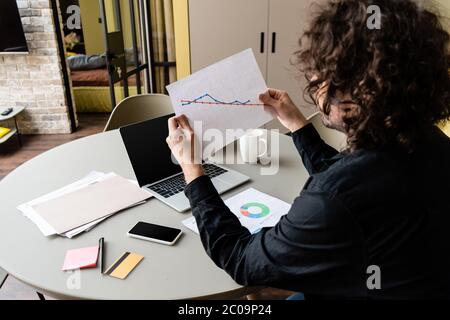 Back view of freelancer holding paper with charts near gadgets and credit card on kitchen table Stock Photo
