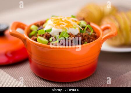 Fresh homemade bowl of chili con carne with beans, sour cream, grated cheddar cheese and green onions. Served with hasselback potatoes on the side. Stock Photo