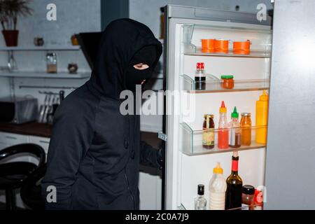 Side view of robber in balaclava opening fridge in kitchen Stock Photo