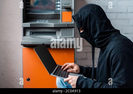 Side view of hacker in mask using laptop with blank screen near atm Stock Photo