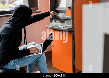 Side view of hacker in mask using atm and laptop with blank screen Stock Photo