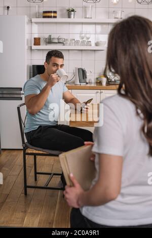 selective focus of mixed race man drinking coffee and looking at woman with book Stock Photo