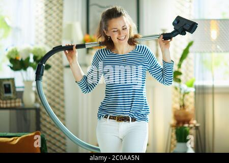 happy young woman in striped t-shirt and white pants with vacuum cleaner in the modern living room in sunny day. Stock Photo