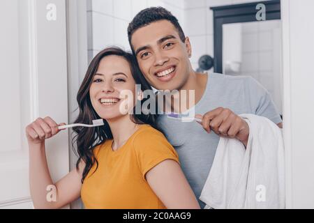happy multiracial couple holding toothbrushes in bathroom 2c0aag9
