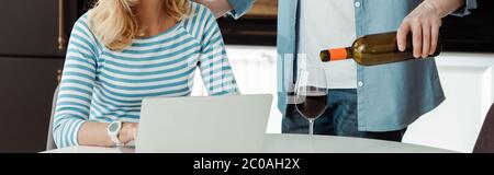 Panoramic shot of man pouring wine near woman using laptop in kitchen Stock Photo