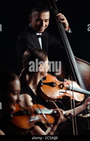 trio of smiling musicians playing on double bass and violins isolated on black Stock Photo