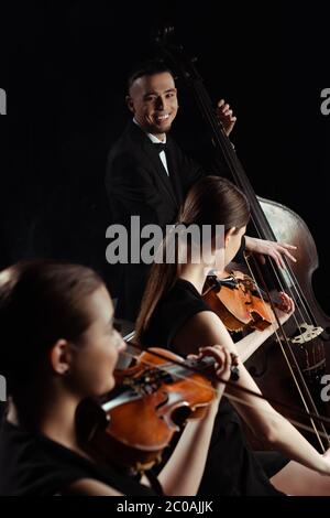 happy professional musicians playing on double bass and violins isolated on black Stock Photo