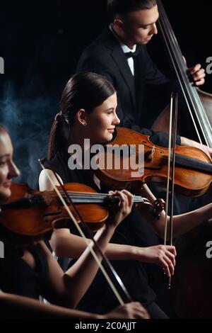 trio of professional musicians playing on violins and contrabass on dark stage with smoke Stock Photo