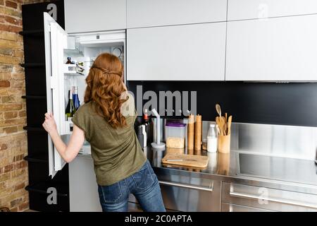 Back view of woman standing near open fridge in kitchen Stock Photo