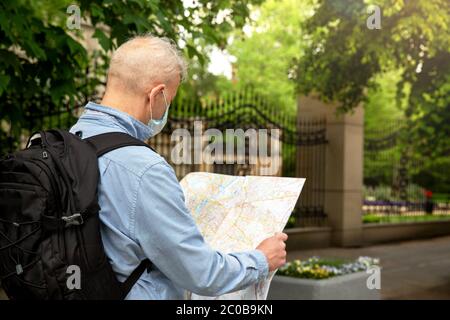 The gray-haired man in a denim shirt and with a backpack looks at the map. Mask on the face. City street. Fence and park are visible. A new travel con Stock Photo