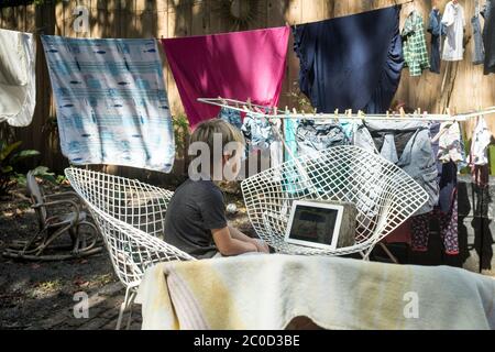 A kindergarten age boy looks at a tablet computer, surrounded by hanging laundry, in the backyard of his home in Miami, Florida Stock Photo
