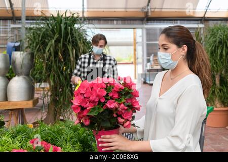 Close up of a focused young woman checking a beautiful plant with an out of focus clerk at the back on an out of focus background. Garden center and s Stock Photo