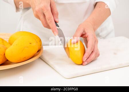Fresh ripe organic yellow mangoes close up. Woman cutting mango on white cutting board, preparing fruit salad Stock Photo