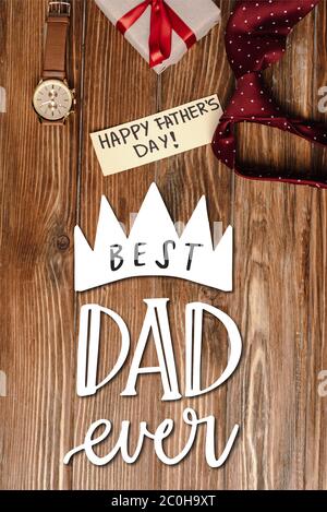 Top view of gift box with red bow, greeting card with lettering happy fathers day and mens red tie and wristwatch on wooden background, best dad ever Stock Photo