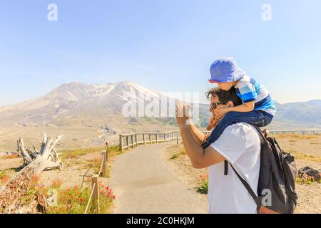 Father is taking pictures on a trace near Mount St. Helens and a little son who sits on fathers shoulders is watching at a screen Stock Photo