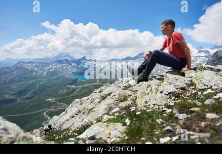 Side view of male tourist sitting on the edge of rocky hill under cloudy sky. Mountaineer admiring the view of mountain valley with grassy hills. Concept of travelling, hiking and tourism.