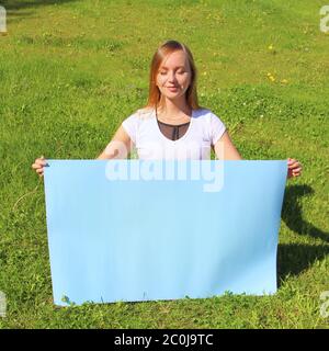 A beautiful long-haired girl on the green grass on the lawn holds an empty blue poster. Layout for design, place for text, copy space. Stock Photo
