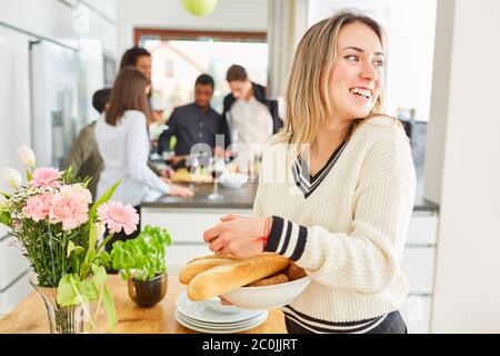 Laughing young woman at table setting with baguette in kitchen in front of a group of friends in shared flat Stock Photo