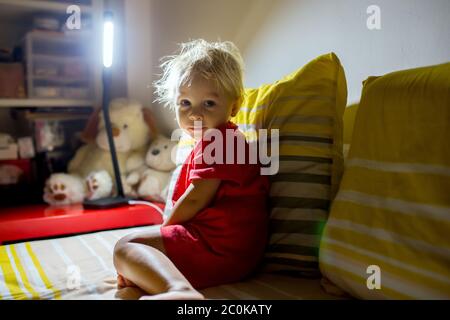 Sweet toddler boy, sitting in bed, prepared for night sleep, nightstnad lamp turned on Stock Photo