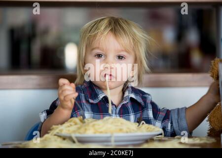 Little baby boy, toddler child, eating spaghetti for lunch and making feeding teddy bear friend, pot with spaghetti and tomatoes on the table Stock Photo
