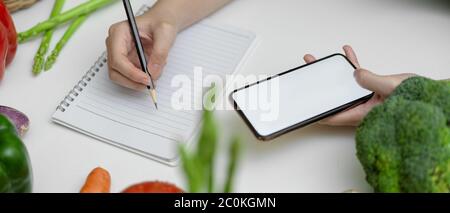 Cropped shot of female noting diet plan while looking information on mock-up smartphone on kitchen table with fresh vegetables Stock Photo