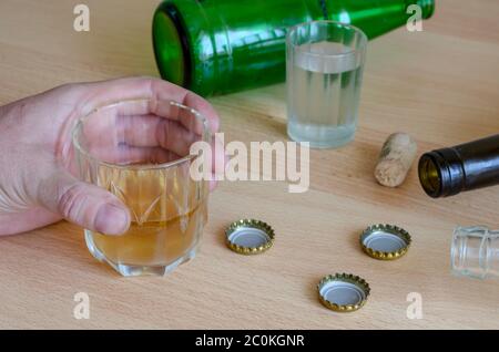 Semi-empty glass cup with beer in a male hand. A glass of vodka, beer corks and empty bottles on the table. Hangover, alcoholism. Social problems. Sel Stock Photo
