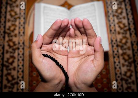 Young Muslim woman praying with Tasbeeh. The Holy Quran is the background, Indoors. Focus on hands. Stock Photo