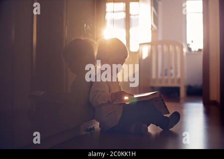 Beautiful toddler blond boy, lying on the floor at home in the hall, reading book, beautiful back light Stock Photo