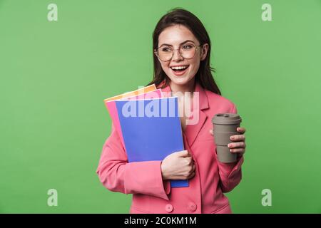 Portrait of a beautiful happy young elegant woman wearing pink jacket standing isolated over green background, drinking takeaway coffee, holding folde Stock Photo